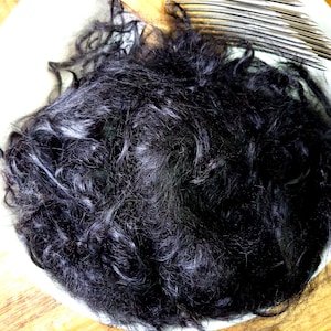 May include: A close-up shot of a white plate holding a large mass of dark, curly hair. The hair appears to be black with some strands of gray. The plate has a simple design and sits on a wooden surface.