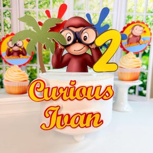 Peut inclure: Un gâteau blanc sur le thème de Curious George, avec une figurine de Curious George avec des jumelles, un chiffre 2 jaune et le nom "Curious Ivan". Comprend des cupcakes et des éléments décoratifs.
