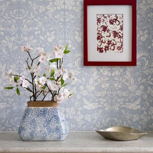 May include: A white ceramic vase with a blue and white floral pattern, filled with a branch of white cherry blossoms, sits on a white marble surface next to a silver metal bowl. A red picture frame with a red and white abstract print hangs on a blue and white floral wallpaper.
