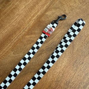 May include: A black and white checkered fabric leash featuring a black metal swivel snap hook. A small rectangular fabric tag is attached near the hook, displaying a red and white graphic of a race car with the text Grand Prix printed on it.