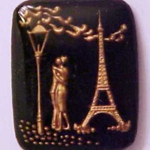 May include: A rectangular black button with a gold embossed design featuring the Eiffel Tower, a lamppost, and a couple embracing. The button has a slightly rounded shape and a glossy finish.