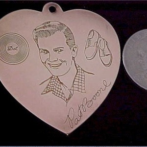 May include: A heart-shaped metal charm with a black and white engraved image of a man with a record and shoes. The charm is signed "Pat Boone".