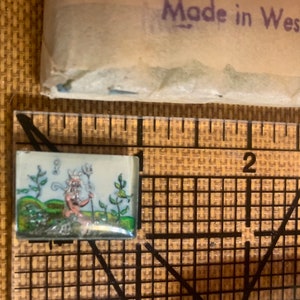 May include: A small, white ceramic tile with a hand-painted image of a whimsical creature with a crown, sitting on a green hill with flowers and plants. The tile is surrounded by a black border.