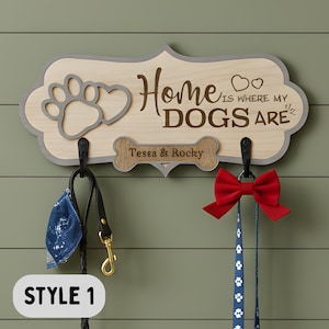 May include: A light-colored wooden wall-mounted leash holder with a gray border, featuring a paw print, two hearts, and the text "Home is where my DOGS are". A bone-shaped plaque displays the names "Tessa & Rocky" above two black metal hooks.