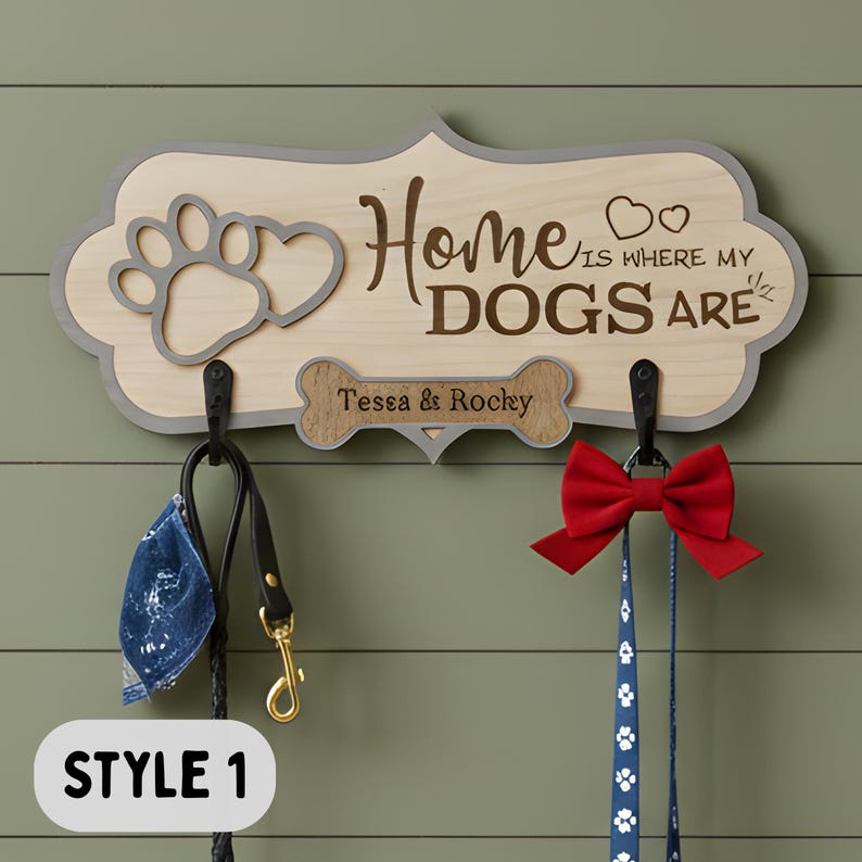 May include: A light-colored wooden wall-mounted leash holder with a gray border, featuring a paw print, two hearts, and the text "Home is where my DOGS are". A bone-shaped plaque displays the names "Tessa & Rocky" above two black metal hooks.