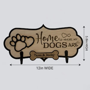 May include: A light wood wall sign with a black border, featuring a paw print, two hearts, and the text "Home IS WHERE MY DOGS ARE". A bone-shaped cutout displays the names "Tessa & Rocky" above two black hooks. It measures 12 inches wide by 5.8 inches high.