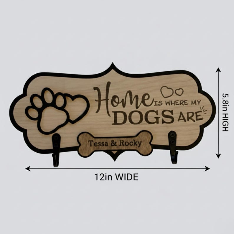 May include: A light wood wall sign with a black border, featuring a paw print, two hearts, and the text "Home IS WHERE MY DOGS ARE". A bone-shaped cutout displays the names "Tessa & Rocky" above two black hooks. It measures 12 inches wide by 5.8 inches high.