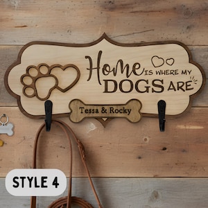 May include: A light-colored wooden wall sign with a dark brown border, featuring the engraved text "Home is where my DOGS are" and a paw print heart design. It includes two black metal hooks and a bone-shaped plaque labeled "Tessa & Rocky".