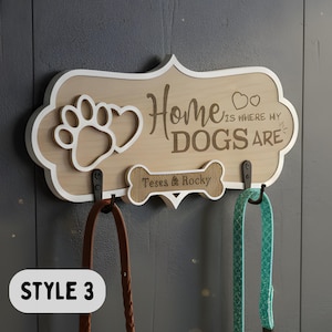 May include: A light wood wall-mounted leash holder with a white border, featuring a cutout paw print and heart, and the text "Home is where my DOGS are". A bone-shaped plaque displays the names "Teses & Rocky" above two black metal hooks.