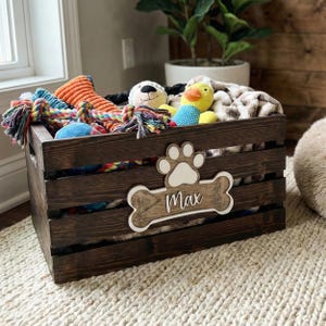 May include: A dark brown wooden crate filled with various colorful pet toys and a plush blanket. The front of the crate features a white paw print and a bone-shaped wooden plaque with the name Max written in white cursive script lettering.