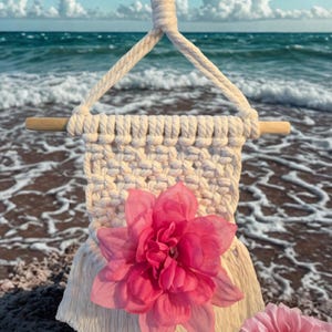 May include: Cream-colored macrame wall hanging with a wooden dowel and a large pink flower attached. The macrame has a woven pattern and fringe at the bottom. A second pink flower is visible in the foreground, with the ocean in the background.