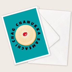Könnte beinhalten: Eine Grußkarte mit türkisfarbenem Hintergrund und den schwarzen Worten "CHAG CHANUKKA SAMEACH!". Die Karte zeigt ein cremefarbenes, rundes Design mit einem roten Punkt in der Mitte, das einem Krapfen ähnelt. Ein weißer Umschlag befindet sich hinter der Karte.