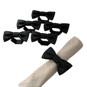 May include: A set of black bow tie napkin rings. The napkin rings are made from a soft, textured fabric. One napkin is rolled and held by a bow tie ring. These rings are a stylish addition to any table setting.