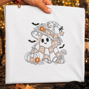 May include: A white t-shirt featuring an embroidered design of a ghost wearing a plaid witch hat. The ghost is surrounded by pumpkins, a mushroom, bats, and small flowers. The embroidery uses black outlines with orange plaid accents.