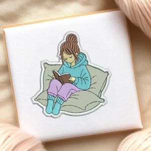 May include: Embroidered image of a person reading a book while sitting on a pillow. The person is wearing a blue hoodie, purple pants, and blue socks. The book is brown, and the pillow is gray. The image is on a white background with a wooden frame.