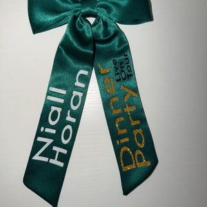 Op de afbeelding: Een donkergroene satijnen strik met twee lange linten. Op het linkerlint staat "Niall Horan" in witte tekst. Op het rechterlint staat "Dinner Party Live On Tour" in goudkleurige glittertekst.
