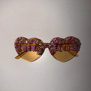Peut inclure: Lunettes en forme de cœur avec un design orné de bijoux roses et violets. Le texte "BEN ELLIS" est affiché sur les verres. Les verres sont de couleur ambre clair.
