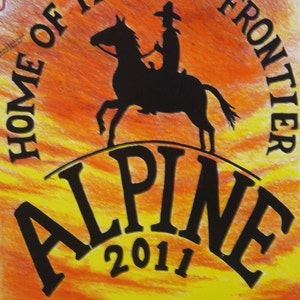 May include: A black and white illustration of a cowboy riding a horse against a sunset background. The text "HOME OF THE LAST FRONTIER ALPINE 2011" is written in a circle around the image.