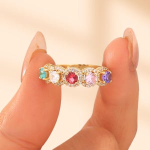 May include: A gold-colored ring featuring a row of colorful gemstones. The gemstones are in shades of teal, white, red, lavender, pink, and purple. The ring is held up by a person's fingers.