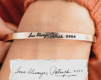 May include: A rose gold-toned metal cuff bracelet featuring a smooth, flat surface engraved with the black cursive text "Love Always, Patrick. XOXO". The bracelet is displayed alongside a torn piece of white paper showing the same handwritten note.