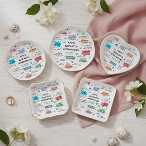 Könnte beinhalten: Eine Sammlung weißer Keramik-Dekoteller in verschiedenen Formen, darunter rund, herzförmig und quadratisch. Jeder Teller trägt den Text "AMY'S BOSS BITCH AFFIRMATIONS" mit farbenfrohen Grafiken und Affirmationen. Goldfarbener Rand.