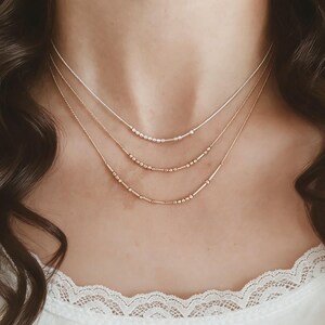 May include: Three layered necklaces featuring delicate metallic chains. Each necklace displays a unique sequence of small round beads and short cylindrical bars, arranged in a pattern that resembles Morse code, resting against the neck.