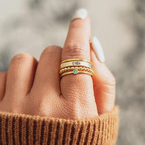 Op de afbeelding: Een stapel gouden ringen aan een vinger. De bovenste ring heeft de naam "Cora" gegraveerd. De tweede ring is een eenvoudige band, en de derde ring heeft kleine gouden kralen. De onderste ring heeft een kleine, ronde, lichtblauwe edelsteen.
