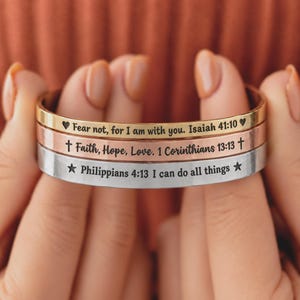 May include: A stack of three metal cuff bracelets in gold, rose gold, and silver tones. Each bracelet features engraved black text and symbols: Fear not, for I am with you. Isaiah 41:10 with hearts, Faith, Hope, Love. 1 Corinthians 13:13 with crosses, and Philippians 4:13 I can do all things with stars.