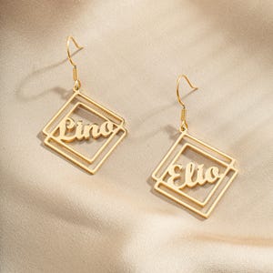 May include: Gold-tone earrings with a square design featuring the names "Lino" and "Elio" in a cursive font.