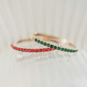 May include: Two gold-toned bangles, one adorned with red gemstones and the other with green gemstones. The bangles are set against a soft, blurred background, highlighting the jewelry's sparkle and design. The gemstones are evenly spaced around the bands.