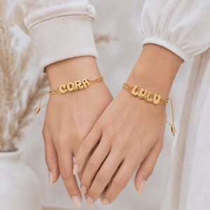 Op de afbeelding: Twee goudkleurige armbanden met bolvormige, afgeronde letterbedels. De ene armband toont de naam CORA en de andere de naam LULU. Beide armbanden zijn gemaakt van dunne, verstelbare goudkleurige kettinkjes met schuifsluitingen.