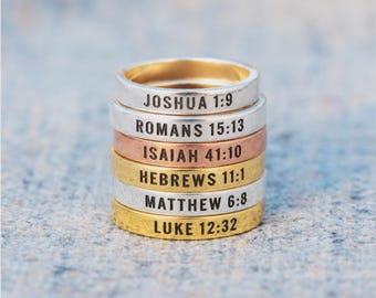 Puede incluir: Una pila de seis anillos de metal en tonos plata, oro y cobre. Cada anillo presenta texto negro estampado: JOSHUA 1:9, ROMANS 15:13, ISAIAH 41:10, HEBREWS 11:1, MATTHEW 6:8 y LUKE 12:32. Las bandas tienen un acabado liso, plano y pulido.