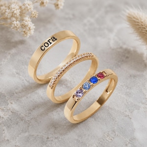 May include: A set of three gold-toned rings. The first ring features the engraved name cora. The second ring is set with a row of small clear stones. The third ring contains four small round stones in purple, light blue, dark blue, and red.