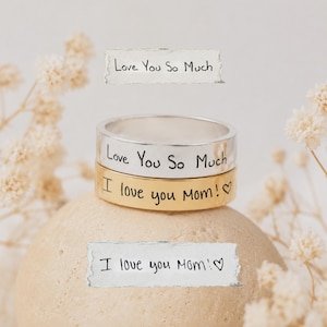 Puede incluir: Dos anillos de metal apilados, uno plateado y otro dorado, cada uno con texto grabado en negro. El anillo plateado tiene la inscripción Love You So Much, mientras que el anillo dorado presenta el texto I love you Mom! con un pequeño corazón.