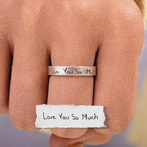 May include: A silver-toned metal ring featuring the engraved text "Love You So Much" in a handwritten style. Below the ring, a small piece of white paper displays the same phrase "Love You So Much" written in black ink, matching the ring's engraving.