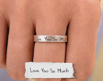Puede incluir: Un anillo de metal plateado con el texto grabado "Love You So Much" en un estilo manuscrito. Debajo del anillo, un pequeño trozo de papel blanco muestra la misma frase "Love You So Much" escrita en tinta negra, a juego con el grabado.