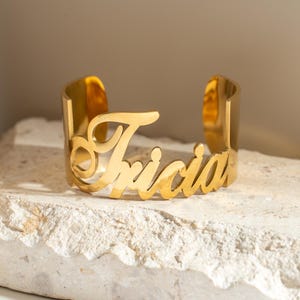 May include: Gold-toned metal cuff bracelet with the name "Tricia" engraved in a cursive font.