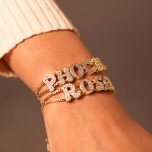 May include: Gold-toned bracelets spelling out "PHOEBE" and "ROSA" in sparkling, clear crystals. The bracelets are layered on a delicate chain, creating a personalized jewelry piece. The bracelets are displayed on a wrist.
