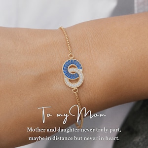 May include: A gold-tone chain bracelet featuring two interlocking circular charms. One circle is set with baguette-cut blue stones, and the other is set with clear pavé stones. Text reads: To my Mom. Mother and daughter never truly part.