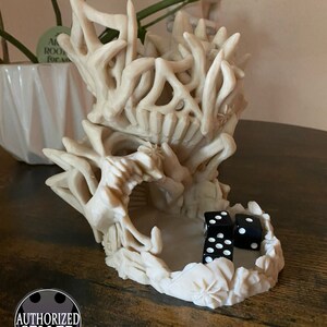 May include: A white 3D printed dragon skull dice tower with three black dice inside. The dice tower is designed to look like a dragon skull with a large open mouth and sharp teeth. The dice tower is sitting on a wooden table.