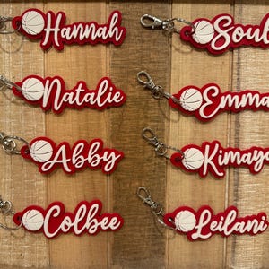 May include: Red and white basketball-themed keychains with names like Hannah, Soul, Natalie, Emma, Abby, Kimaya, Colbee, and Leilani. Each keychain features a basketball graphic and a silver clasp.