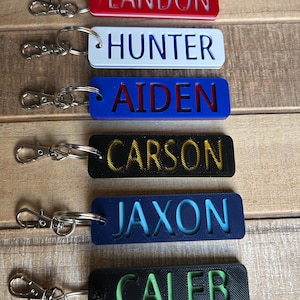 May include: A collection of personalized keychains in various colors, each with a name engraved. The keychains are rectangular with rounded corners and feature names like "LANDON", "HUNTER", and "AIDEN". Each keychain has a silver-colored clasp.