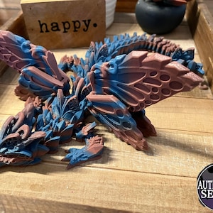 May include: A 3D printed dragon sculpture with intricate details. The dragon is painted in shades of blue and pink, with a butterfly-like wing design. The sculpture is resting on a wooden surface.