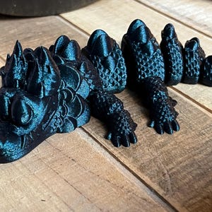 May include: A dark teal dragon figurine with detailed scales and claws. The dragon is posed on a wooden surface, showcasing its articulated body and intricate design. The figurine's head is prominently displayed, with sharp features.