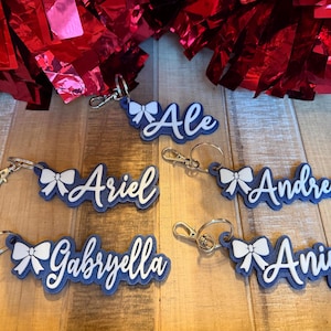 May include: Keychains with names: Ale, Ariel, Andrea, Gabriella, and Aniya. Each keychain features a white bow design and a name in a cursive font, set against a blue background. They have silver key rings and clasps. Red pom-poms are in the background.