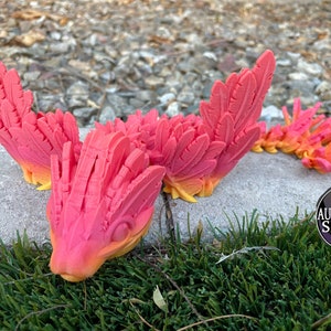 May include: A bright pink and orange 3D printed dragon with feathery wings. The dragon is lying on its belly with its head resting on the ground. The dragon is made of a smooth, plastic-like material.