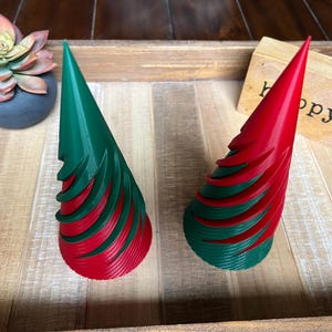 May include: Two red and green spiral Christmas tree ornaments. The ornaments are made of plastic and have a cone shape. They are sitting on a wooden surface.