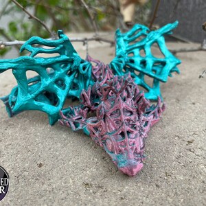 May include: A 3D printed dragon sculpture with a teal blue body and pink wings. The dragon is lying on its side with its head resting on the ground.