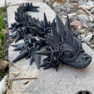 May include: A black 3D printed model of a spiky, reptilian creature with a detailed head and body. The creature is posed in a crawling position with its head raised.