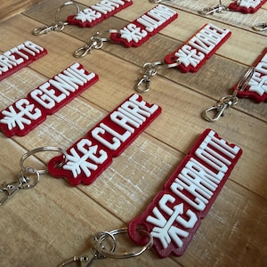 May include: A collection of rectangular red keychains with white raised lettering featuring various names including Loretta, Gennie, Claire, Charlotte, Juliana, Izabel, and Demi. Each keychain includes a silver metal split ring and a lobster clasp.